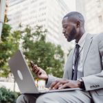 Businessman in a suit using a laptop and smartphone in an urban setting, blending technology with business. — Creating Memora