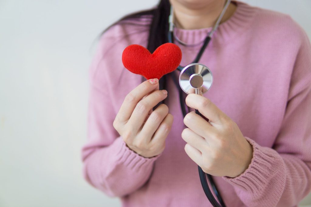 Unrecognizable female in pink sweater with stethoscope on neck standing on white background with red heart in hand in dayligh