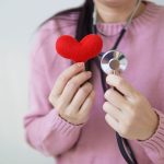 Unrecognizable female in pink sweater with stethoscope on neck standing on white background with red heart in hand in dayligh