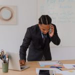 Professional businessman multitasking with phone and laptop at office desk, surrounded by documents. — Gary Guglielmo: Your G