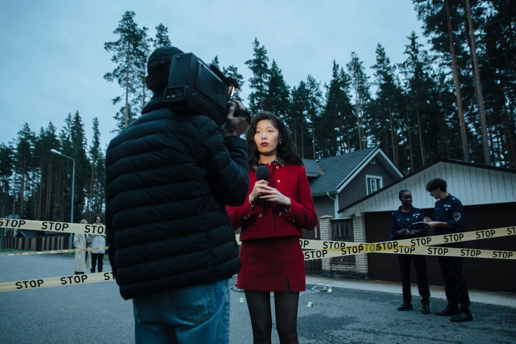A female journalist reports live from an outdoor crime scene, flanked by police officers and media personnel. — Get Your Favo