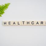 Healthcare word spelled with Scrabble tiles next to a green fern leaf on a white background. — Keith Myers at the Helm of Mor