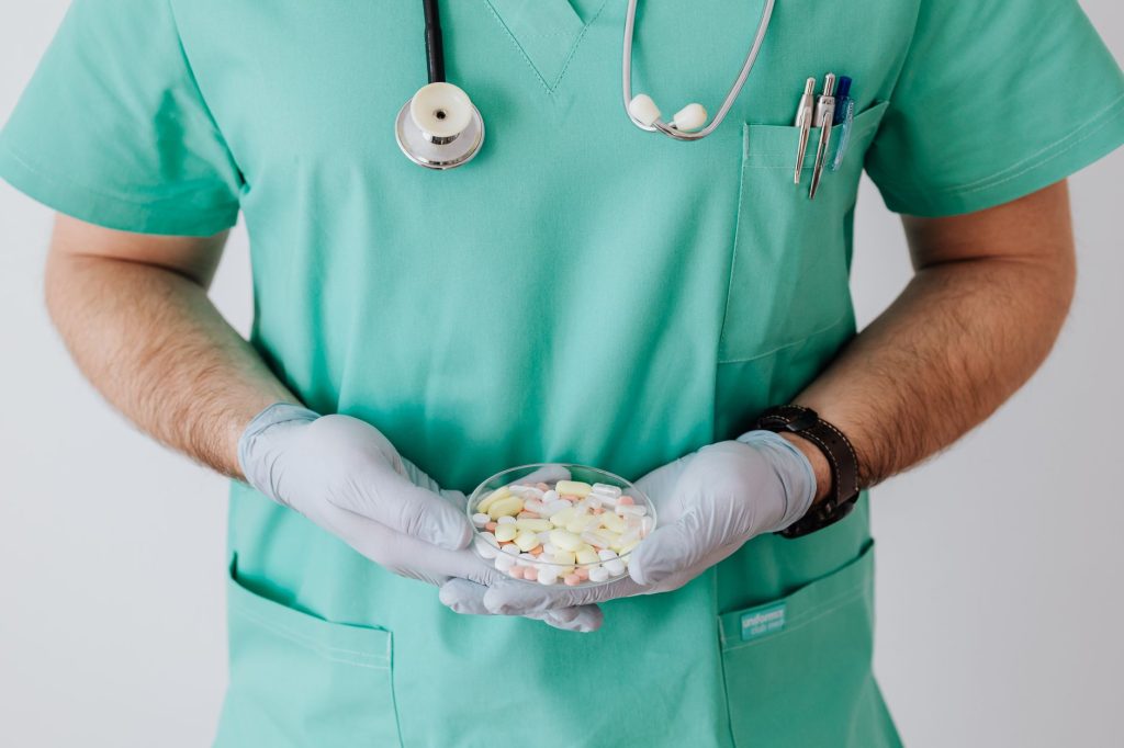 Unrecognizable male doctor wearing medical uniform and latex gloves holding Petri dish filled with assorted drugs while stand