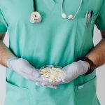 Unrecognizable male doctor wearing medical uniform and latex gloves holding Petri dish filled with assorted drugs while stand