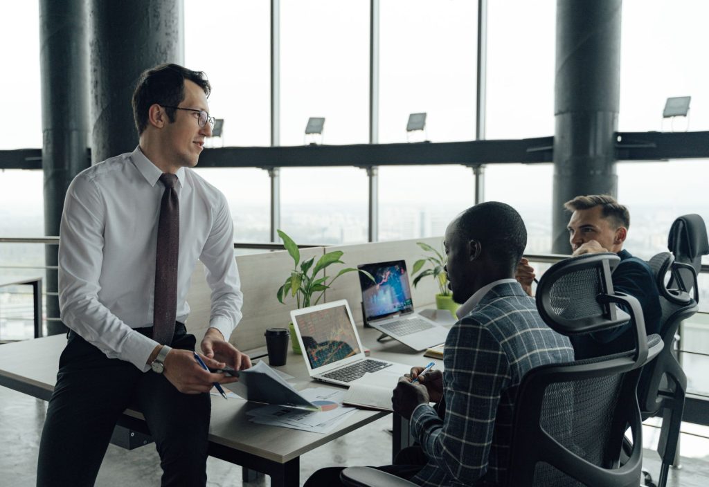 Three men engaged in a business meeting in a modern office setting with laptops and documents. — Tina Wellman: Revolutionizin