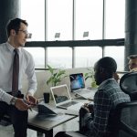 Three men engaged in a business meeting in a modern office setting with laptops and documents. — Tina Wellman: Revolutionizin