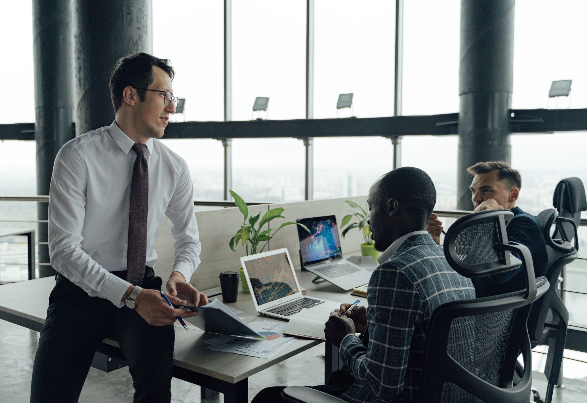 Three men engaged in a business meeting in a modern office setting with laptops and documents. — Tina Wellman: Revolutionizin